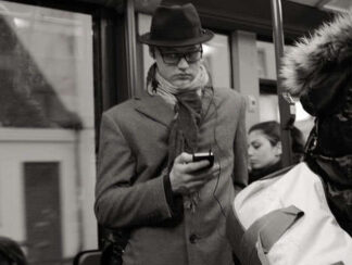 Persona en un autobús, con gabardina y sombrero, mirando su teléfono móvil con una mano y con la otra, agarrado a los apoyos del autobús.