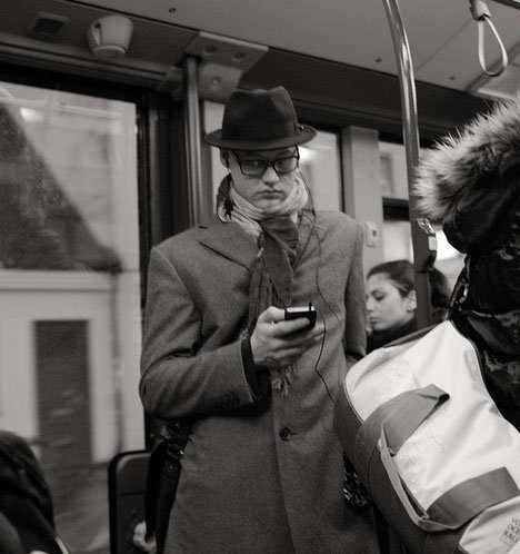 Persona en un autobús, con gabardina y sombrero, mirando su teléfono móvil con una mano y con la otra, agarrado a los apoyos del autobús.