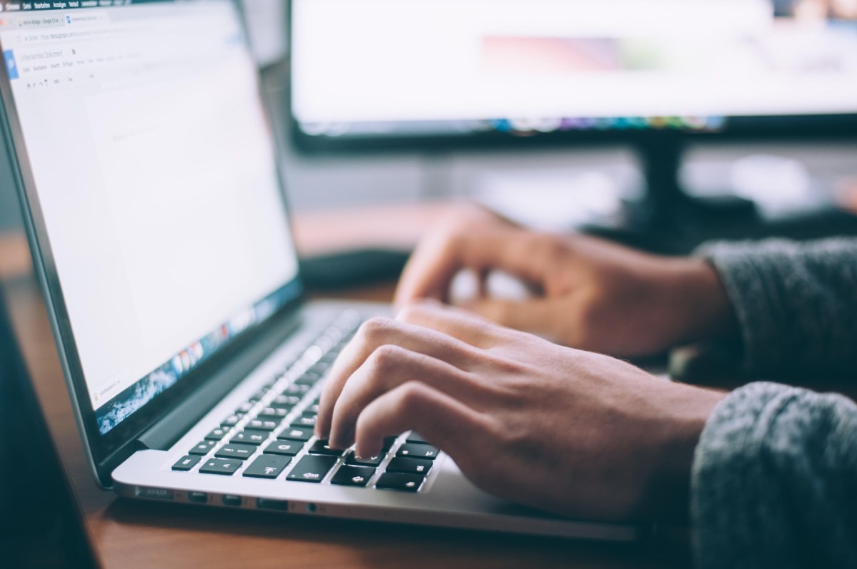 Persona escribiendo en una computadora portátil con un documento abierto en la pantalla, en un entorno de trabajo con un monitor adicional al fondo."